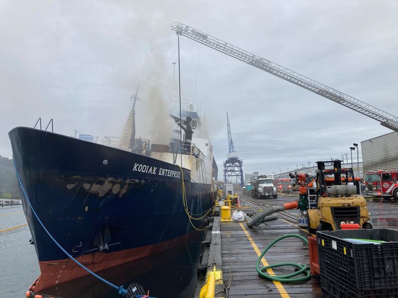 Smoke comes from a vessel next to a dock. A firetruck ladder extends over the boat.